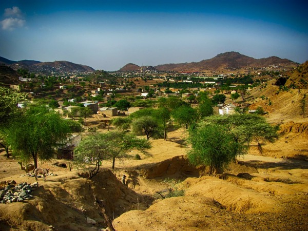 Cette photo montre un paysage de Ghinda en Erythrée : des collines de terre sèche et des maisons entre des arbres verts, sous un ciel bleu.