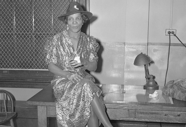Cette photographie en noir et blanc montre Stephanie St. Clair, assise sur une table, les jambes croisées. Elle porte une robe aux manches mi-courtes et s"arrêtant sous les genoux, et un chapeau.