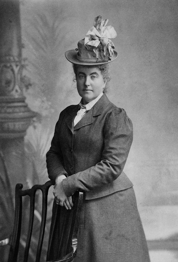 Cette photographie en noir et blanc est un portrait en pied de Fanny Bullock Workman. Debout, les mains sur le dossier d'une chaise, elle porte une jupe et une veste, ainsi qu'un chapeau avec un noeud de ruban.