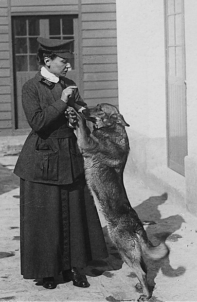 Cette photographie en noir et blanc montre Nicole Girard-Mangin avec sa chienne Dun (pour Verdun). Nicole est débout, en jupe et veste sombre avec une casquette et joue avec sa chienne, debout sur ses pattes arrières.